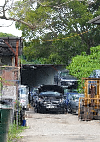 An outdoor area filled with cars, some stacked on top of each other, indicating a car junkyard or repair facility. Vehicles appear in various states of disassembly, with an open hood on the black car in the middle. A forklift is visible near the right, surrounded by green foliage and trees.